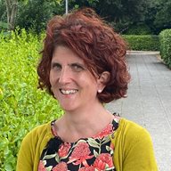A close up of Clara Strauss wearing a yellow cardigan and flowery top in front of a path and green bush