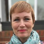 A head shot of a woman called Lara Tozer, stood in front of a wall smiling at the camera wearing a scarf