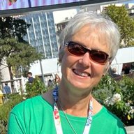 A photo of Val Jenkins wearing a lanyard, sunglasses and a green top in front of a building with glass and trees