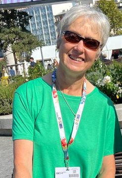 A photo of Val Jenkins wearing a lanyard, sunglasses and a green top in front of a building with glass and trees