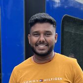 a global health alumni student named Abiman wearing a mustard t shirt in front of a blue train smiling