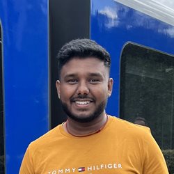 a global health alumni student named Abiman wearing a mustard t shirt in front of a blue train smiling