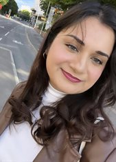 A photo of a girl with brown hair and white top, titled and looking to the right of the camera with pavement in the background