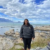 A global health alumni student named Syra stood on rocks with the sea and blue sky in the background