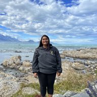 A global health alumni student named Syra stood on rocks with the sea and blue sky in the background