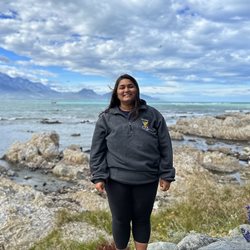 A global health alumni student named Syra stood on rocks with the sea and blue sky in the background