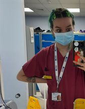 A student called Willow Neal taking a photo on her phone in front of a mirror wearing purple scrubs with a blue facemask. Blue lockers are in the background, showing she is about to go in to a hospital setting