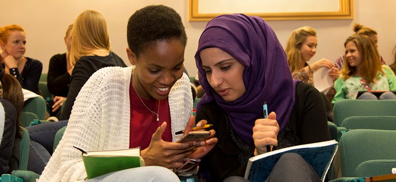 Students sitting in a lecture theatre, smiling and looking at a phone